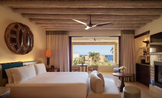 Luxurious hotel room with white‑linen bed, wooden‑beam ceiling, seating area, balcony view of palm trees and ocean.