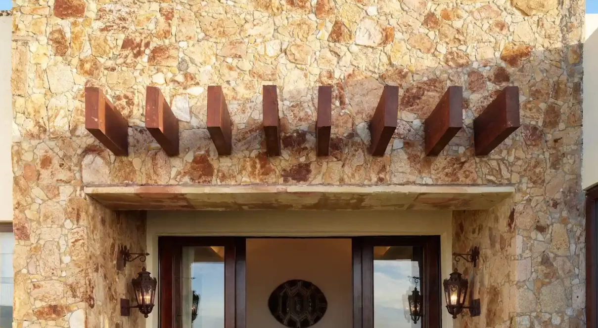Stone‑facade entrance with wooden beams, glass doors, lanterns, and a cozy seating area inside.
