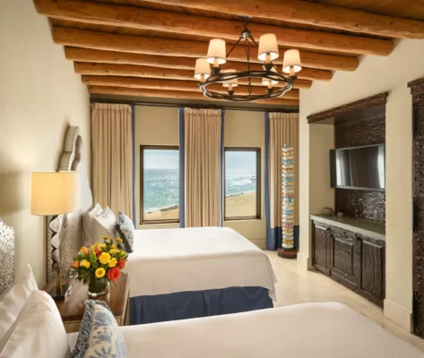 Ocean-view room with two queen beds, wood beams, chandelier, and coastal decor.