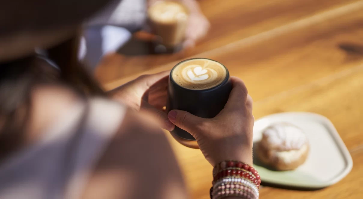 Outdoor coffee break with latte art, beaded bracelets, and pastry on a wooden table.