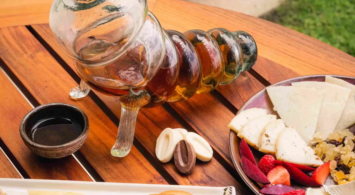 Outdoor table with fruit, cheese, and horn shaped glass decanter in a scenic garden setting.