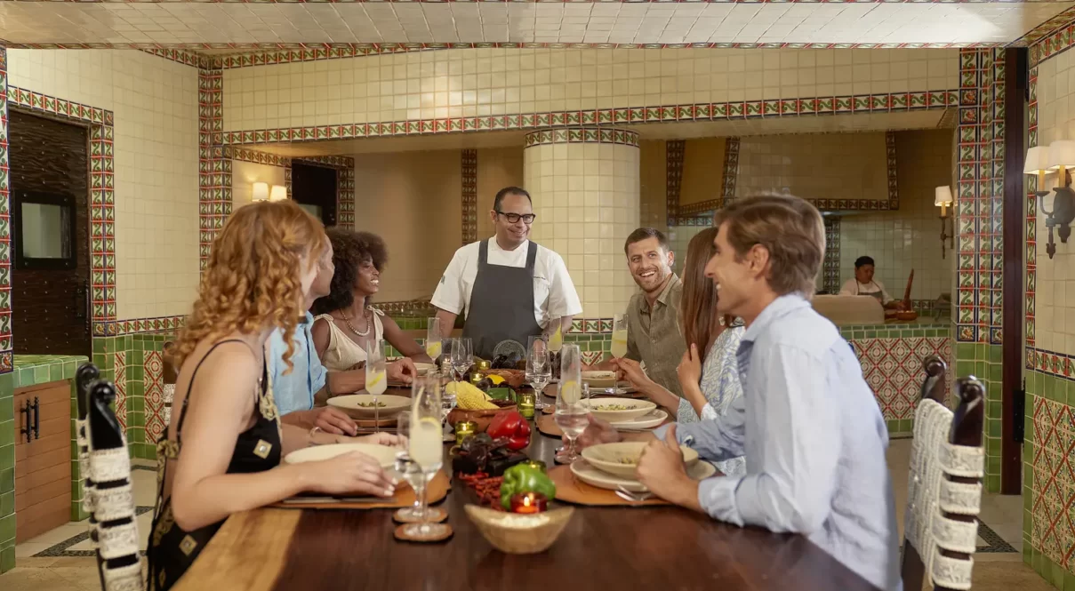 Chef engaging with guests at a festive table filled with fresh produce in a vibrant restaurant setting.