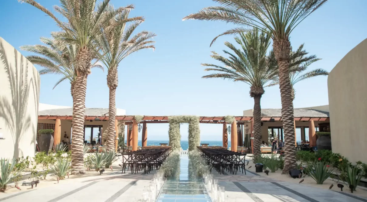 Glass aisle with candles, palm trees, and ocean-view arch in a tropical outdoor wedding setting.